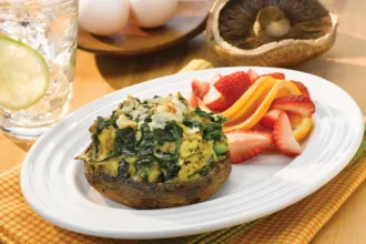 Plate on a placemat with a herbed spinach quiche Portabella mushroom cap and sliced strawberries and oranges, surrounded by a glass of water, small wooden plate with two eggs, and an uncooked Portabella cap.