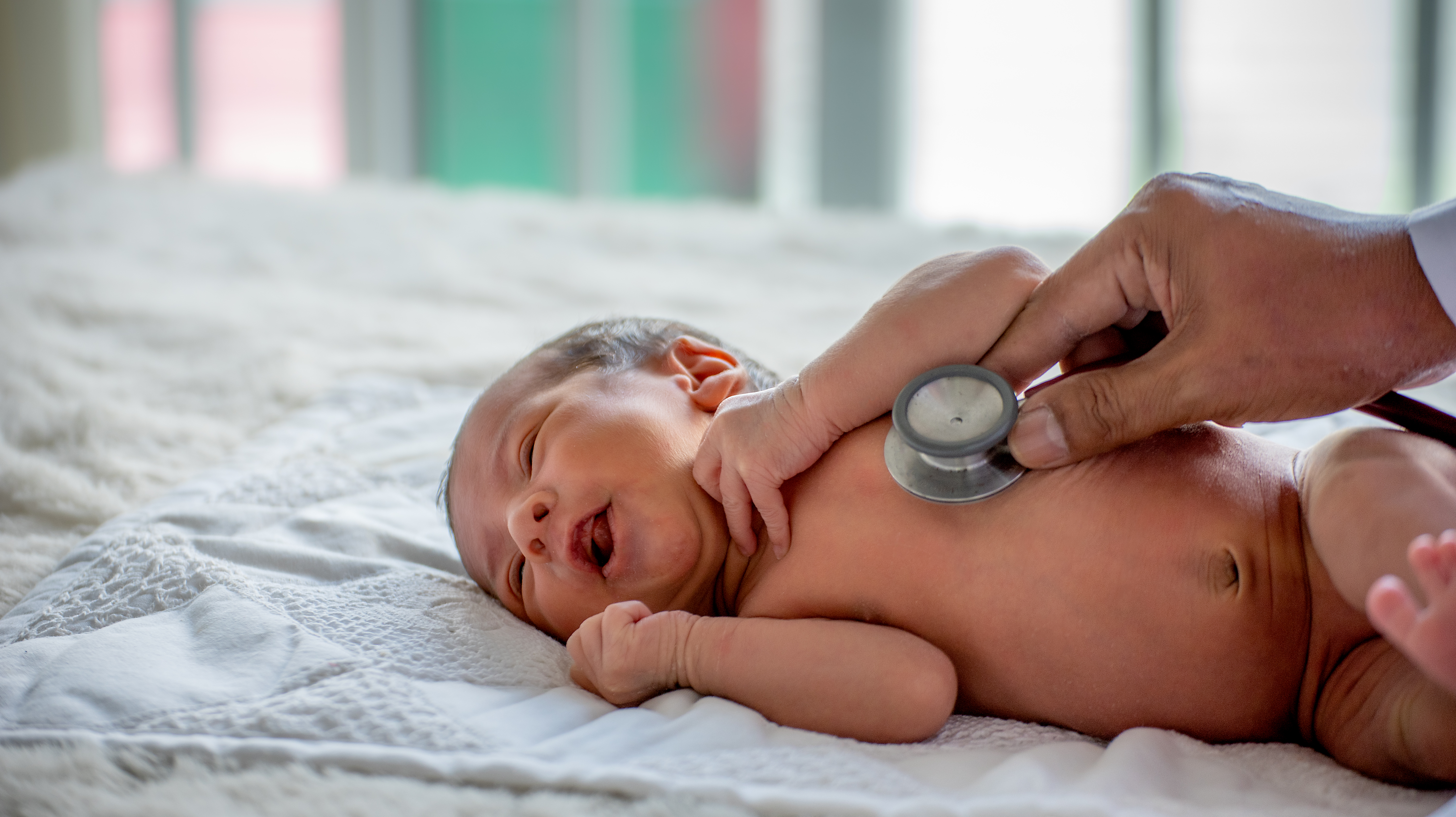 Newborn baby on a bed with hand holding a stethoscope to the baby's chest.