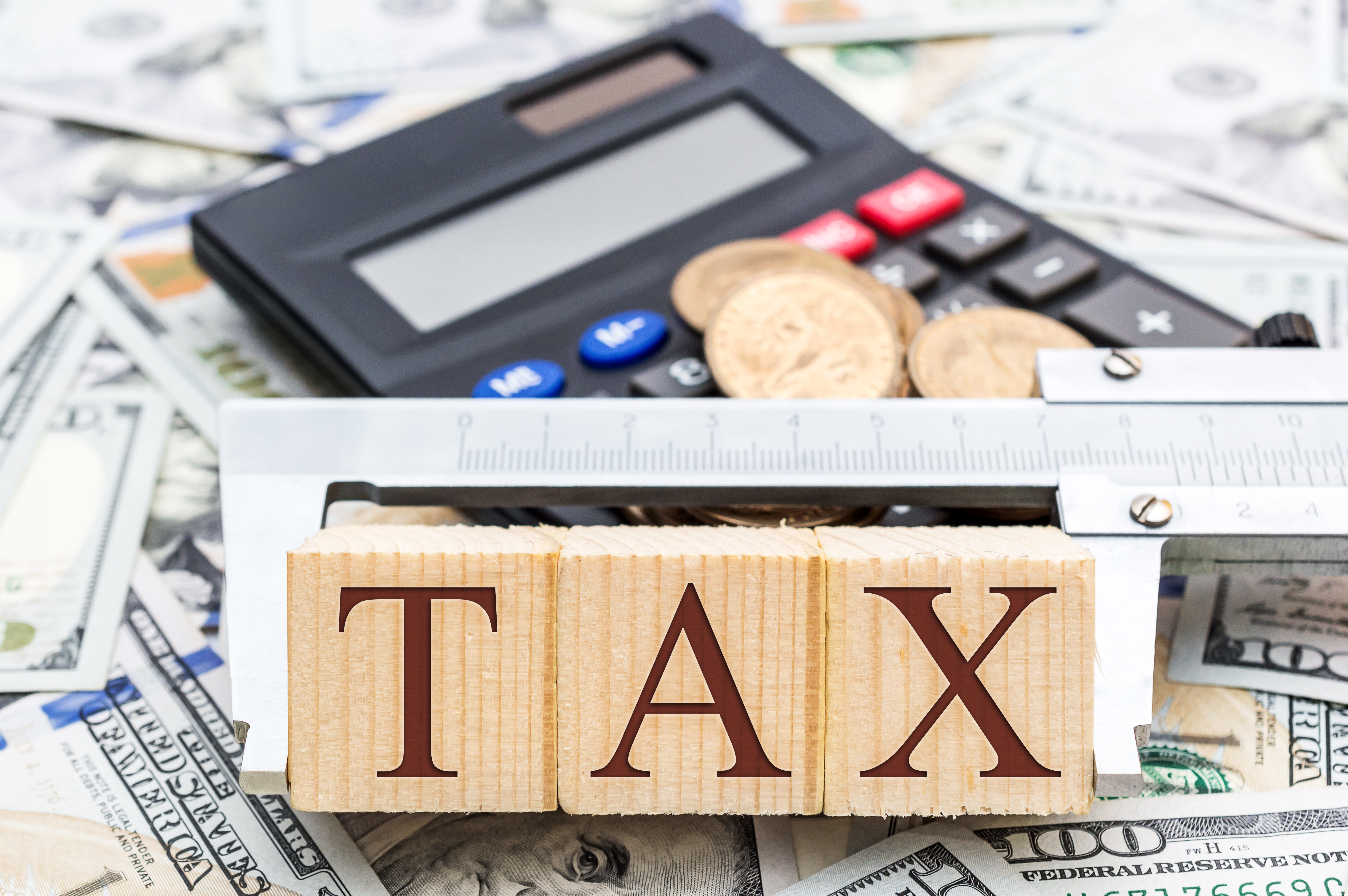 Word "tax" on wooden blocks held together with a caliper tool. A calculator and assorted coins and currency are in the background.