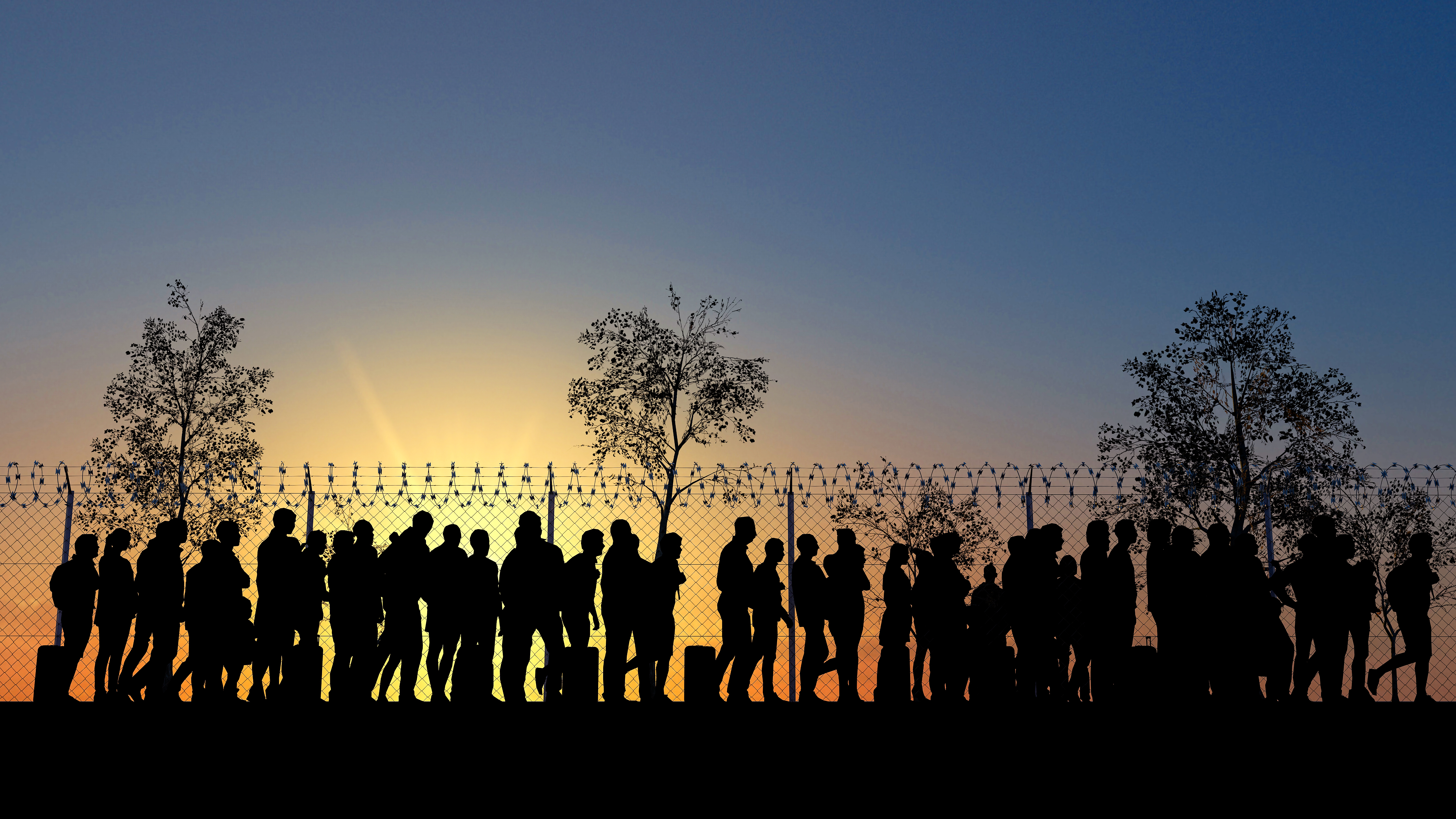 Silhouetted line of people at a fence topped with razor wire at sunrise or sunset.