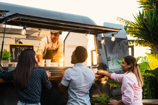 Three adults stand at the window of a food truck, and a man inside the food truck smiles at them and wears an apron.