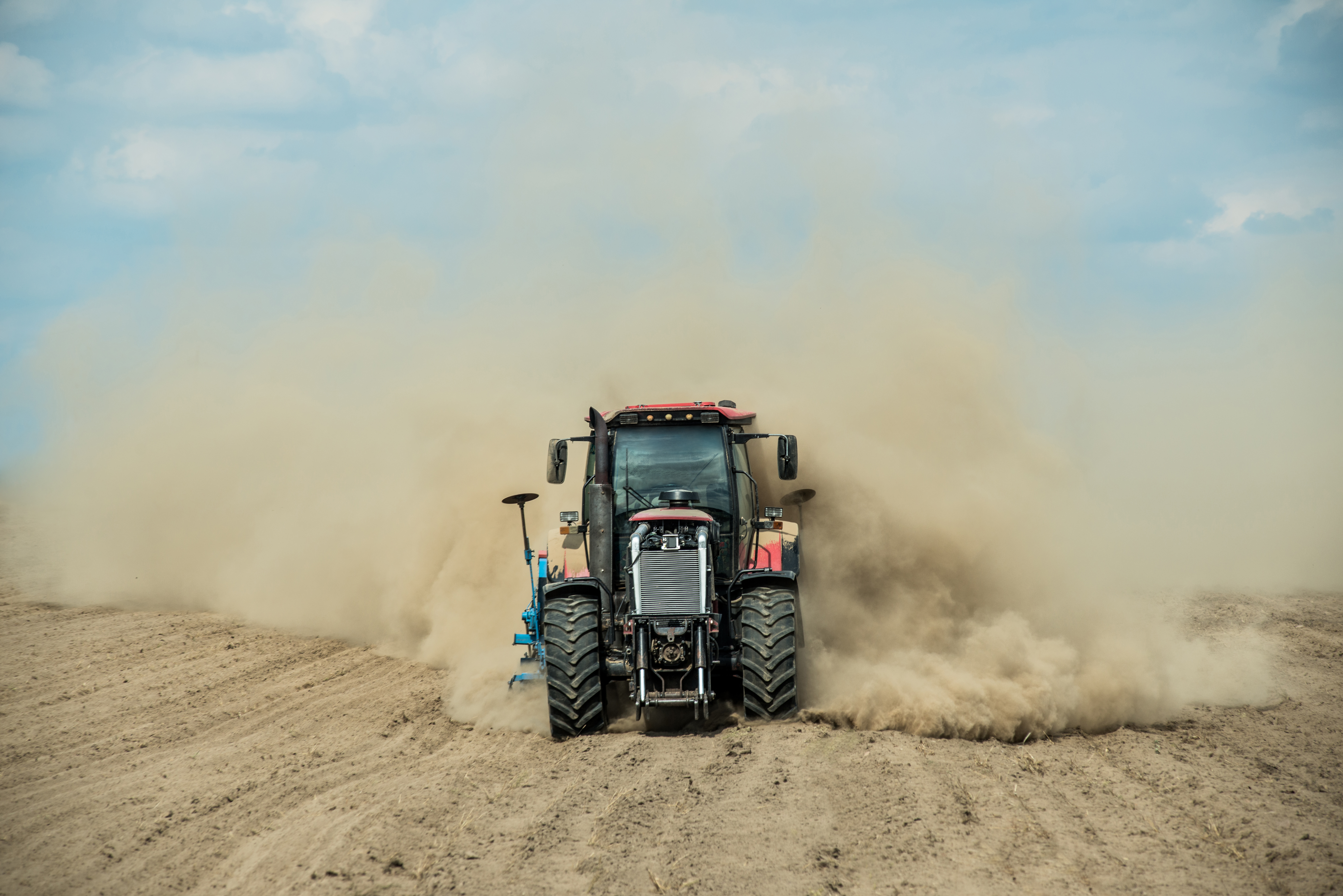 Farm tractor driving through bare dirt creating a dust cloud.
