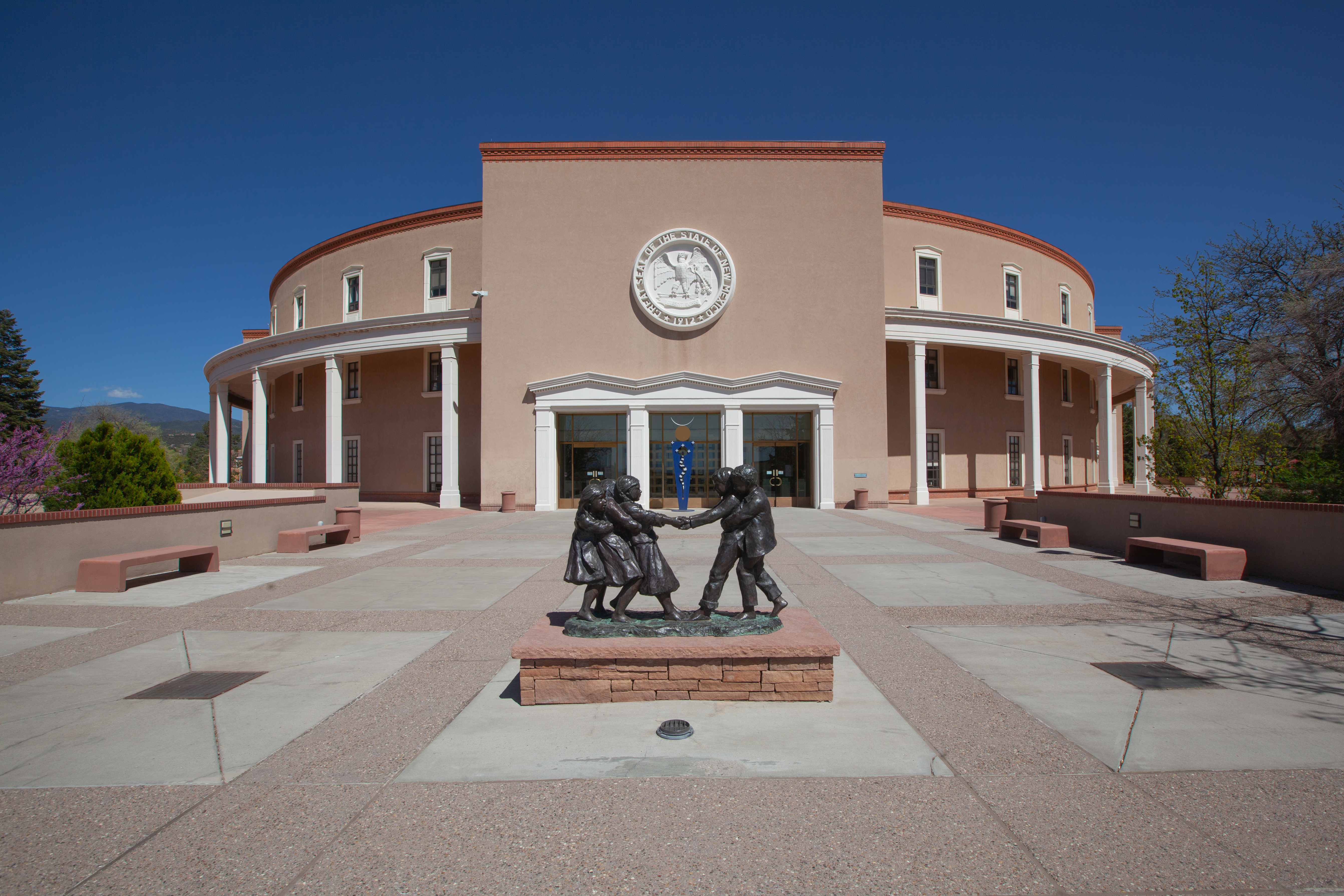 Entrance to the New Mexico State Capitol building. A statue of people is in a plaza area in front of the building.