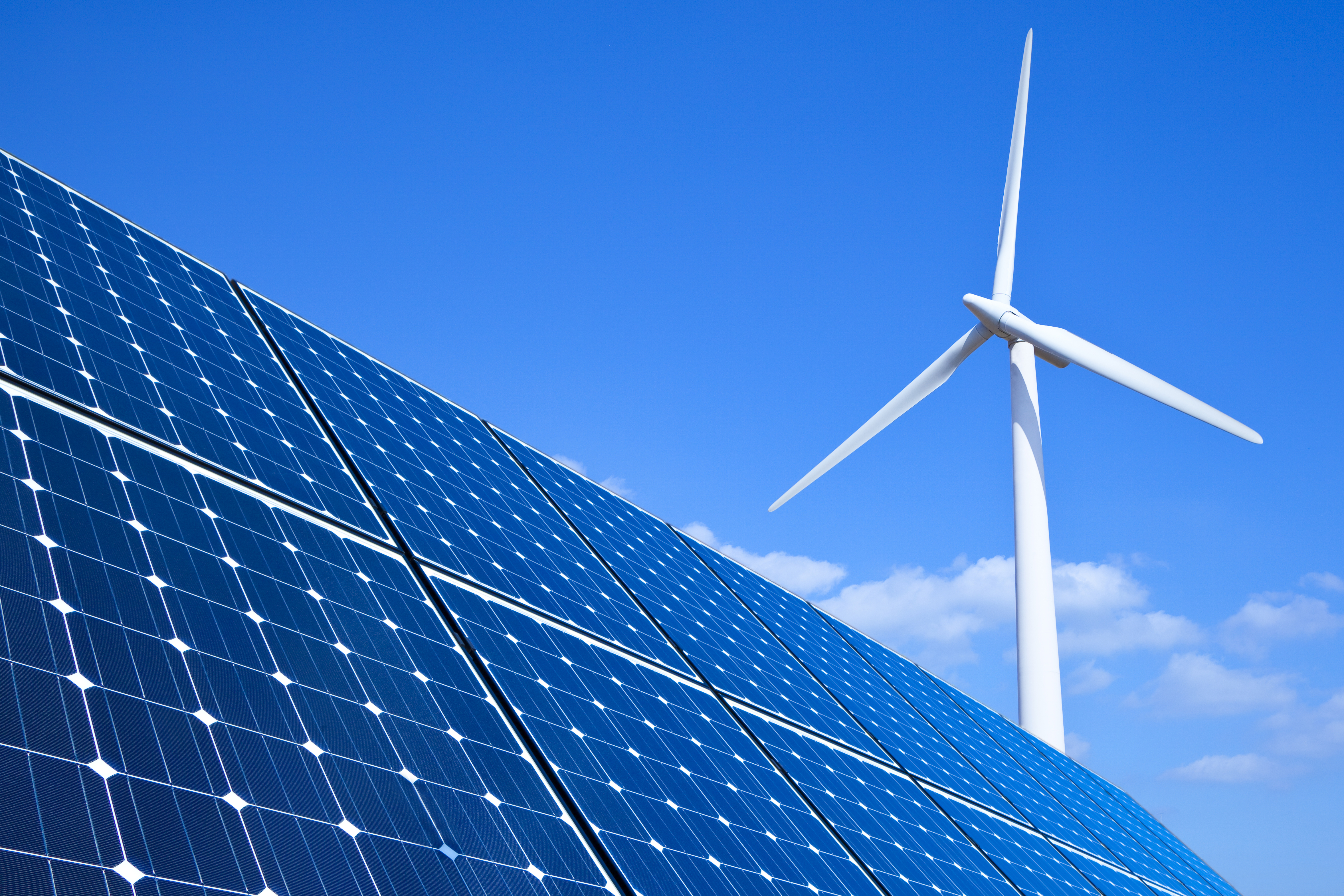 Solar panels in the foreground, with a wind turbine in the background, against a blue sky with a few clouds.