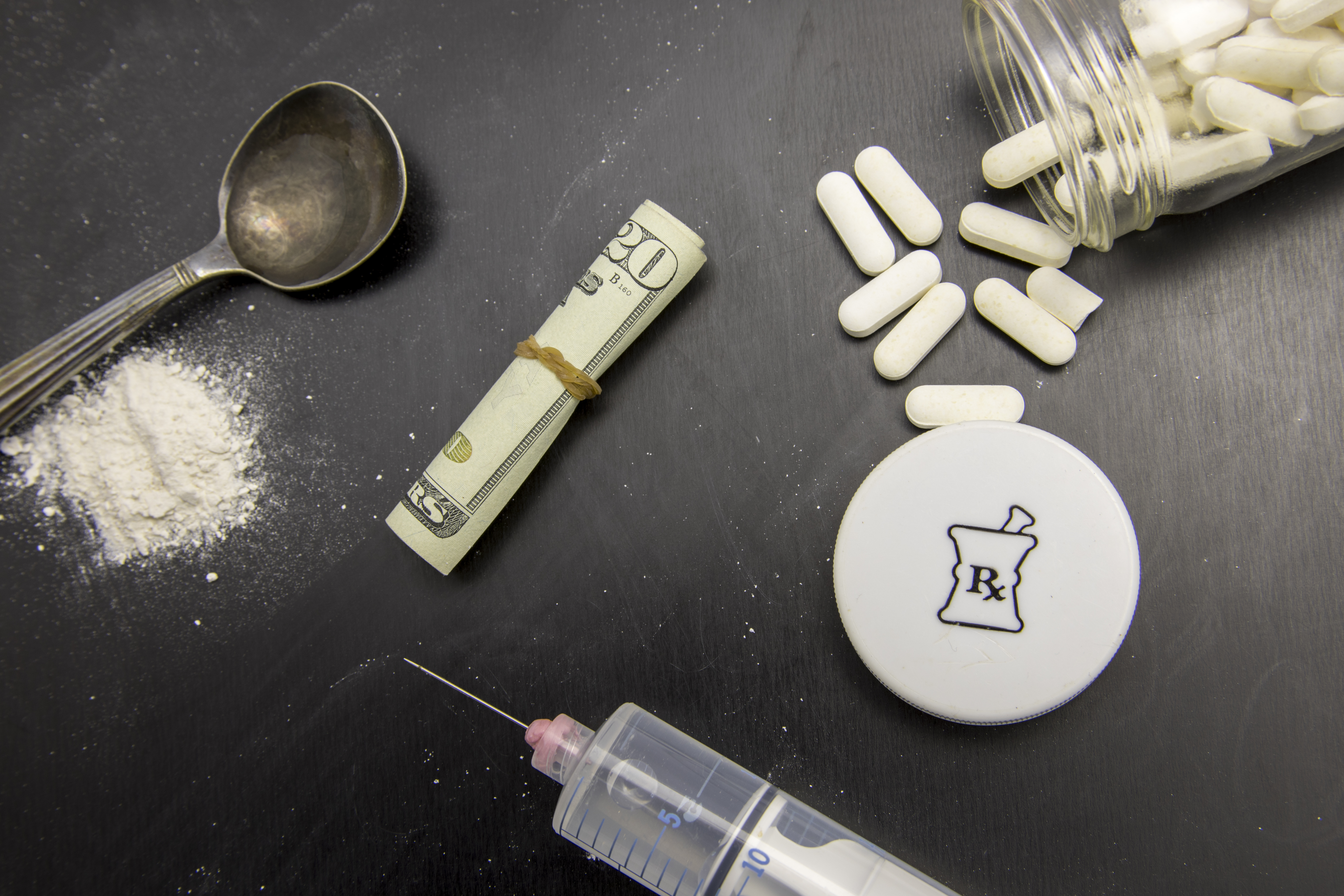 Overhead view of white powder next to a spoon, hypodermic syringe and needle, a rolled up United States 20 dollar bill with rubber band around it, and an open pill bottle with the contents partially spilled.