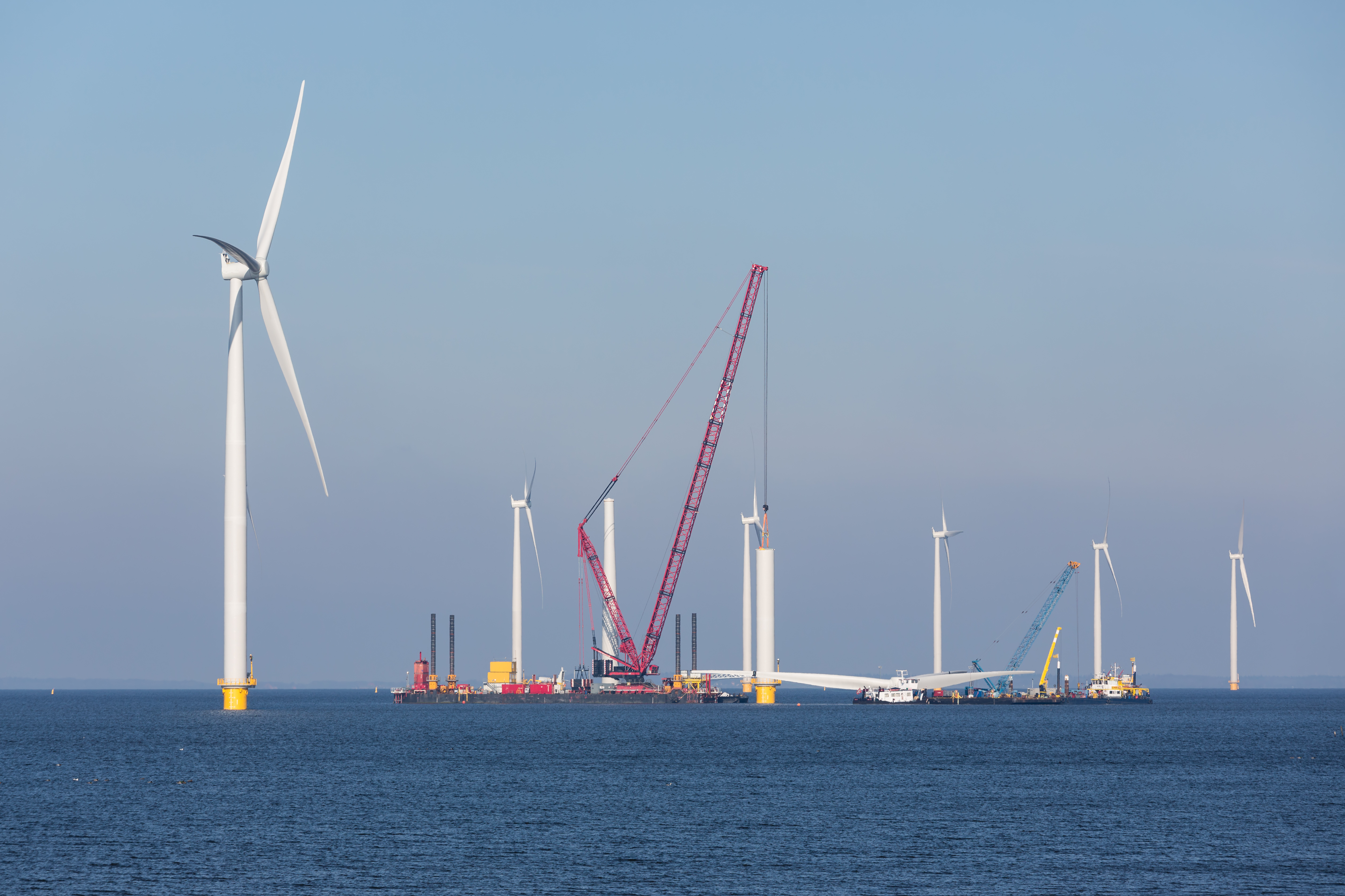 Wind turbines under construction in the ocean. A crane ship is in place to lift a blade assembly.