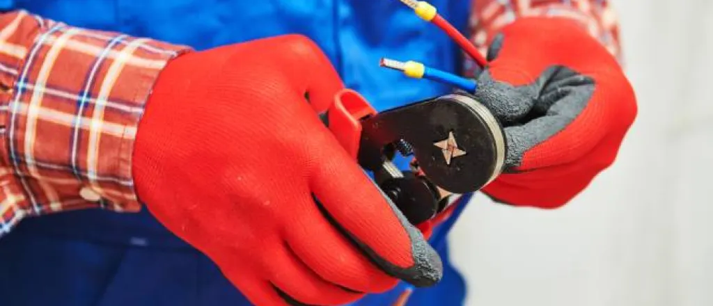 An electrician wearing red working gloves crimping multiple electrical wires with a hand crimping tool.