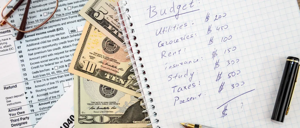 Notebook with a pen on a page showing a personal budget. Underneath are paper currency and tax forms. Eyeglasses are nearby