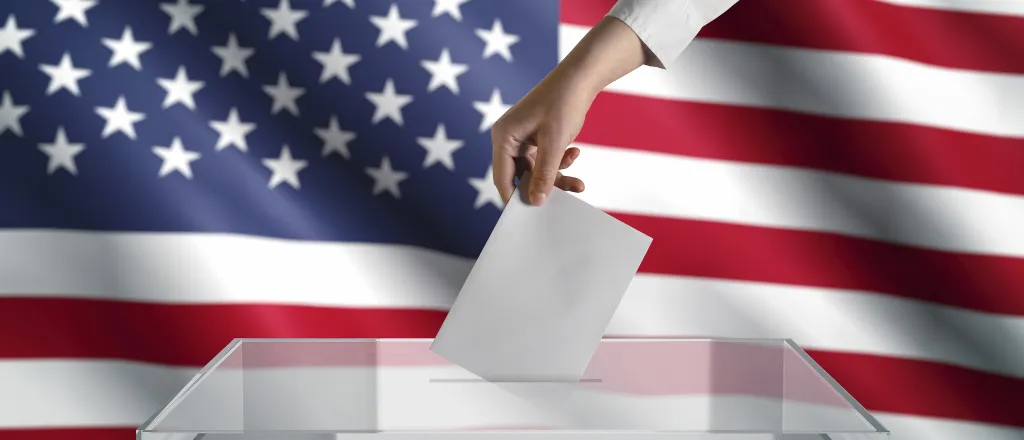 PROMO HIRES Miscellaneous - Election Vote Ballot Flag - iStock - sefa ozel Hand placing a piece of pager into a clear ballot box in front of the United States flag