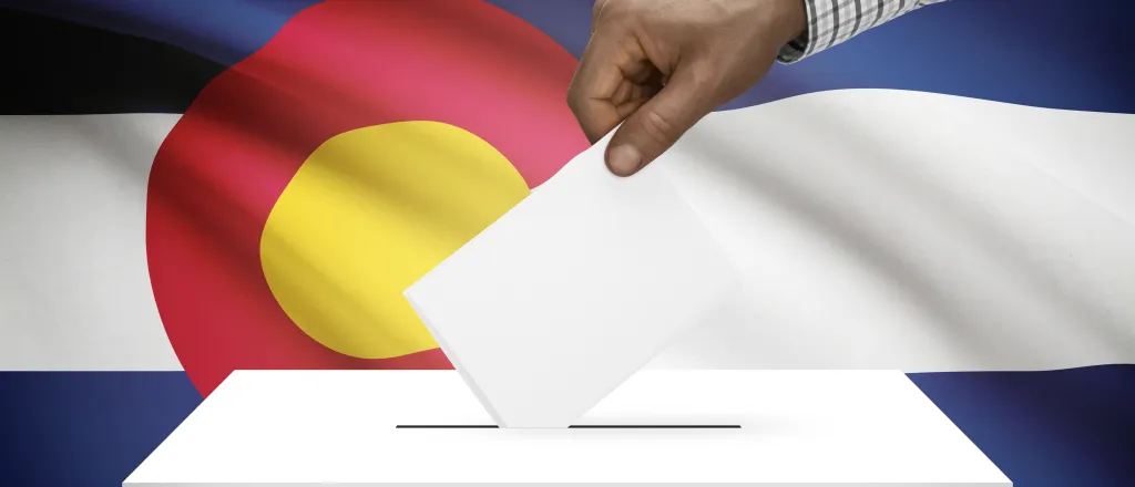 Hand inserting a piece of paper into a ballot box in front of the Colorado flag.