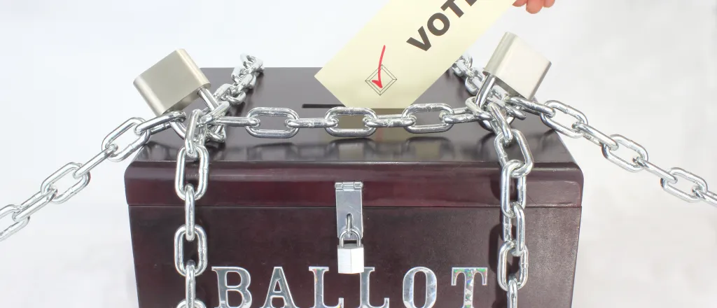 Hand inserting a piece of paper labeled "vote" into a wood box labeled "Ballot Box." The box is secured by chains