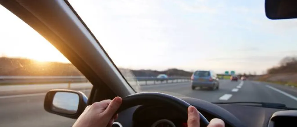 A person grips the steering wheel in their car as they drive on the highway with cars ahead of them and the sun setting.