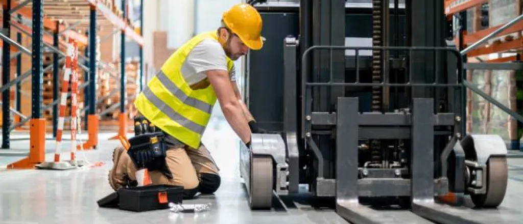 A mechanic kneeling next to a forklift in a warehouse as he makes repairs with a tool kit by his side.