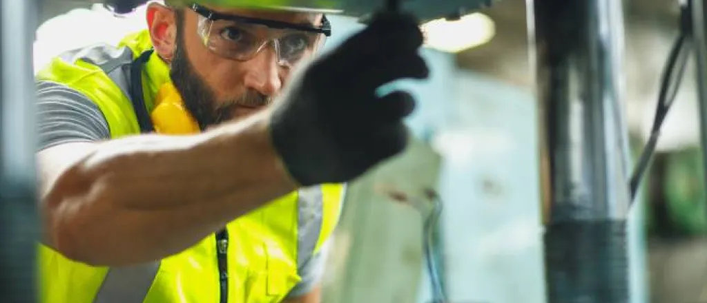 A man in high-visibility gear, safety glasses, and gloves turning hardware on a piece of industrial equipment.