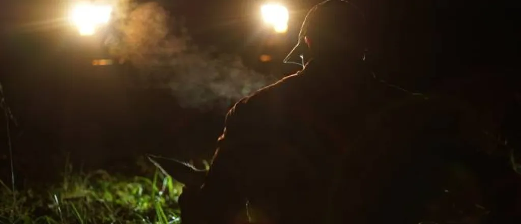 A hunter wearing a cap silhouetted in front of his truck's headlights as he carries the body of a deer toward his vehicle.