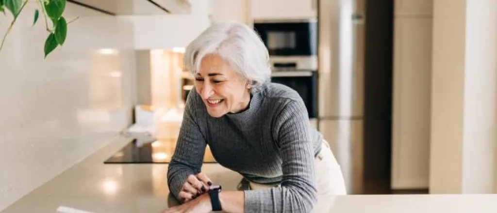 An older woman smiling as she leans over the kitchen counter to talk to her smart home assistant and adjust her smart watch.