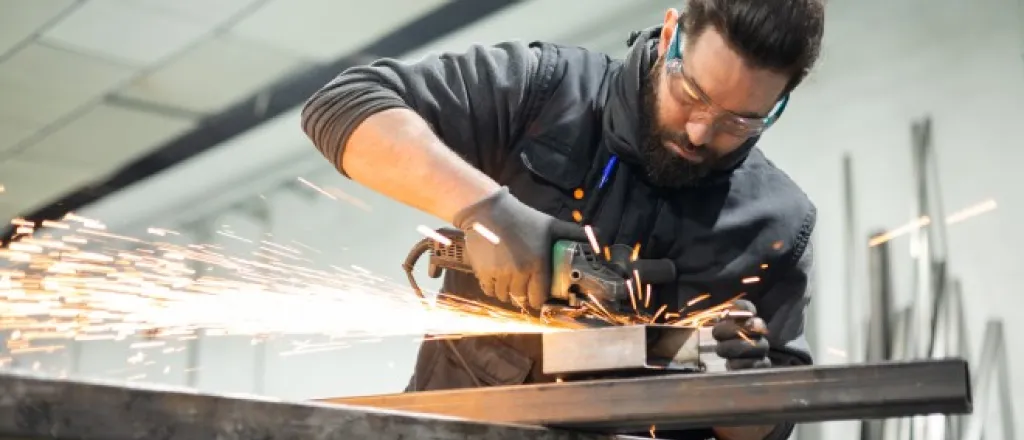 A man working hard in a machine shop with a glass grinder machine and sparks flying. He's wearing proper PPE.