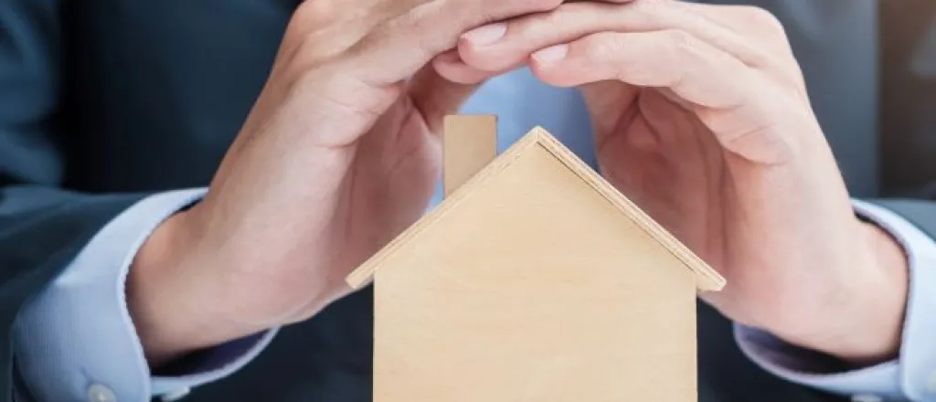 A man in a suit sitting at a table and covering the roof of a small wooden home model with both hands.