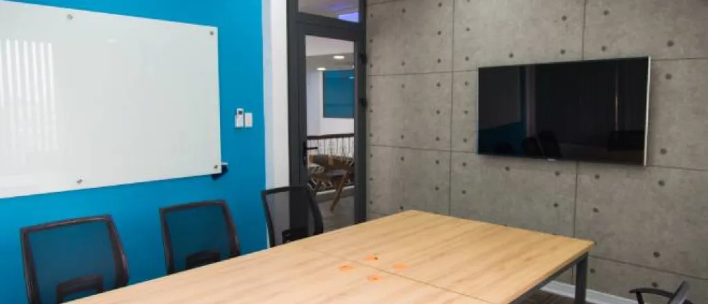 A wooden table in a conference room surrounded by black office chairs. A whiteboard hangs on a blue wall.