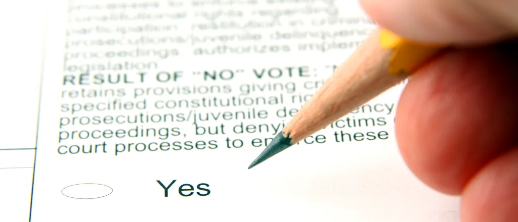 Fingers holding a pencil over an election ballot showing yes and no options