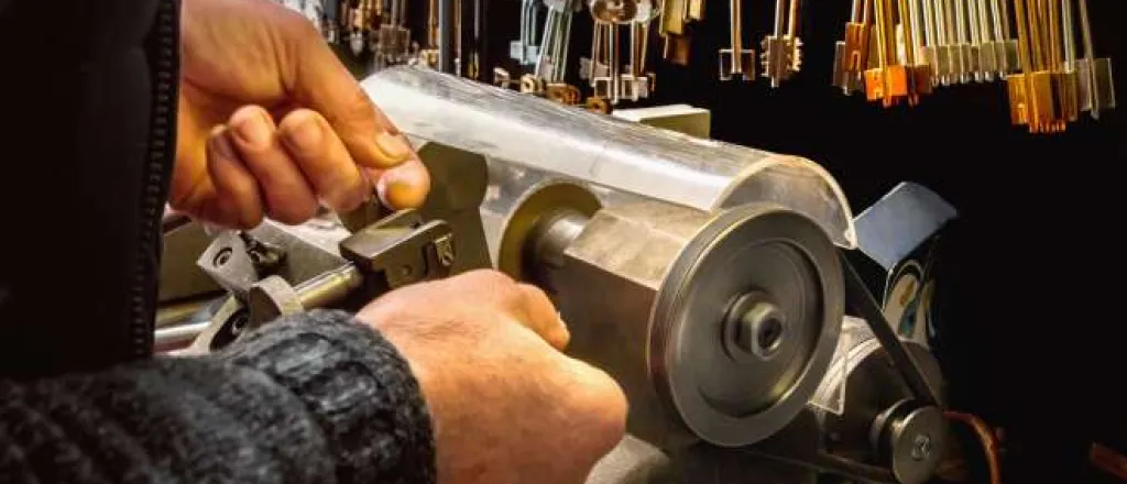 A locksmith using a key copying machine to make a duplicate key. Key blanks are hanging on a wall in front of them.