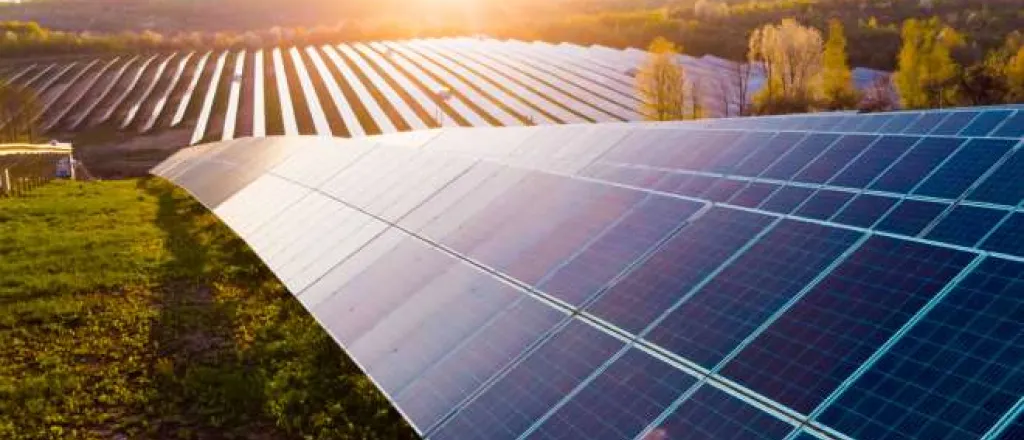 A row of solar panels on a solar farm perched on a hill. The sun is setting in the background, casting a bright glow.