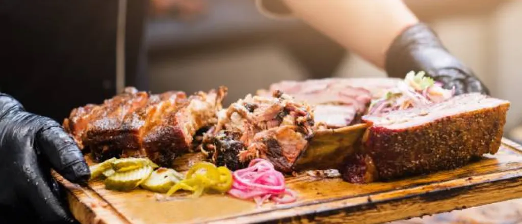 A man with gloved black hands holding a wooden tray of meat and vegetables in a professional restaurant setting.