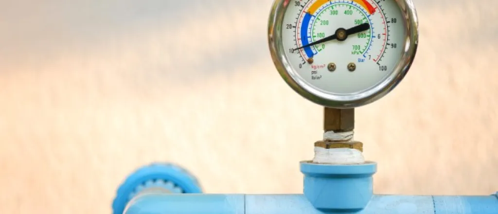 A pressure gauge with metal casing attached to a bright blue pipeline connected to an off-white wall.