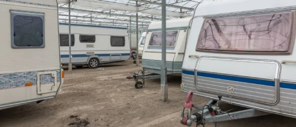 An RV storage facility contains numerous caravans parked on a dirt lot. They are covered by a large tarp acting as a roof.