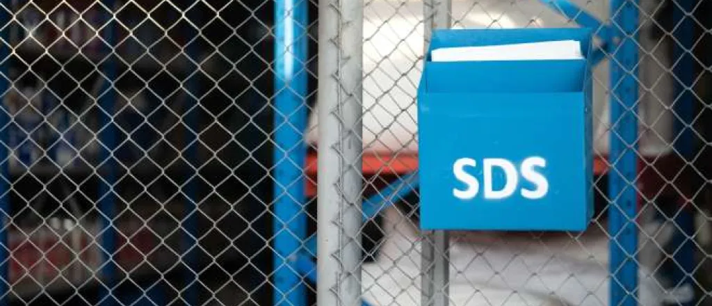 A blue metal box labeled "SDS" hangs on a chain-link fence and holds documents. Storage containers are behind the fence.