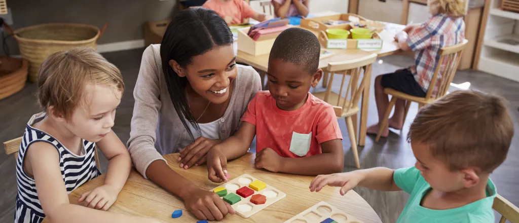 Young children and a care giver at tables engaged in educational activities.