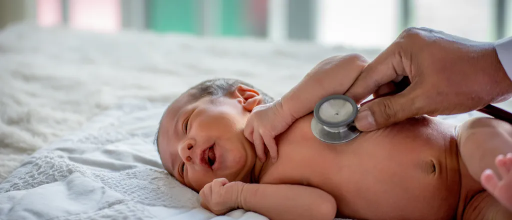 Newborn baby on a bed with hand holding a stethoscope to the baby's chest.