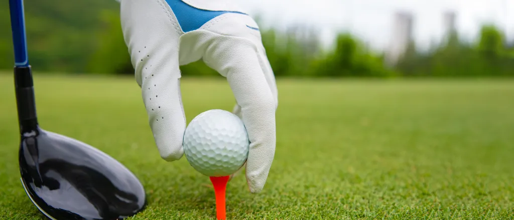 Close up of a gloved hand setting a golf ball on a tee. A golf club is near the tee.