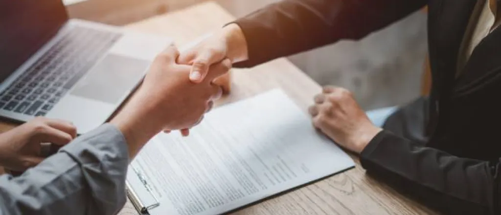 A buyer and a seller sitting at a table together shaking hands after reaching a deal. The contract is sitting on the table.