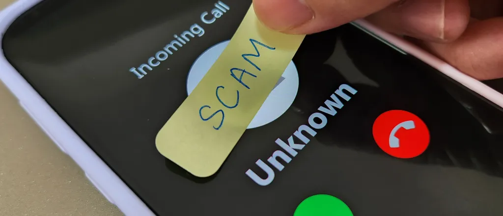 Smartphone showing an incoming call from an unknown number. Fingers are placing a hand-written label with the word "scam" over the top.