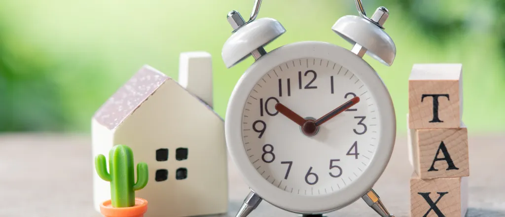 Analog alarm clock flanked by a stack of wooden blocks with the word "tax" and a small house figurine.