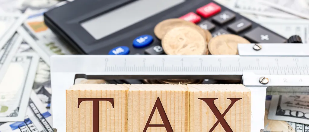 Word "tax" on wooden blocks held together with a caliper tool. A calculator and assorted coins and currency are in the background.