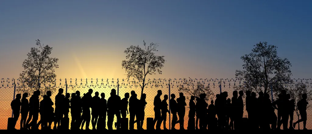 Silhouetted line of people at a fence topped with razor wire at sunrise or sunset.