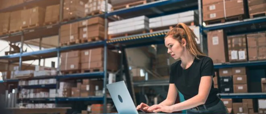 A woman with a ponytail wearing a black shirt standing in front of a table using a laptop in a warehouse. A woman with a ponytail wearing a black shirt standing in front of a table using a laptop in a warehouse.