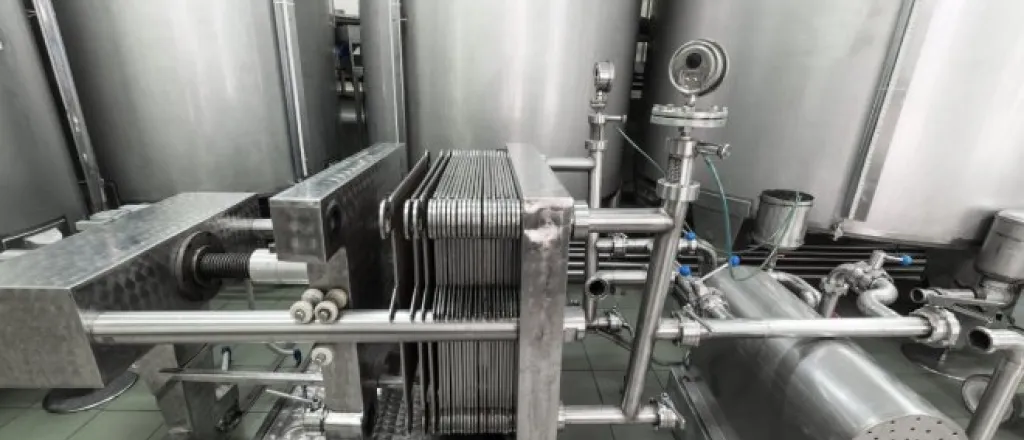 A large stainless steel automatic filter press sits in a room with large stainless steel storage containers.