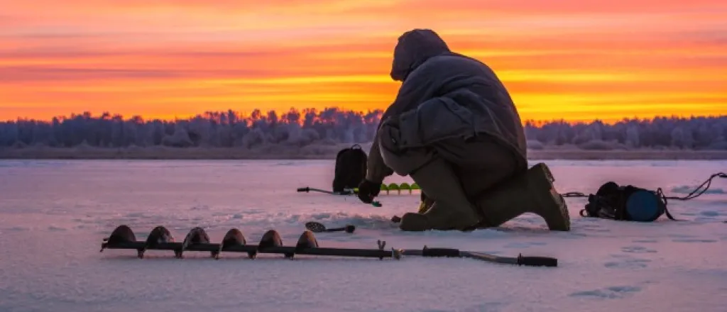 A man kneeling on an icy snow-filled field preparing an ice fishing hole with an auger drill attachment on the ground.