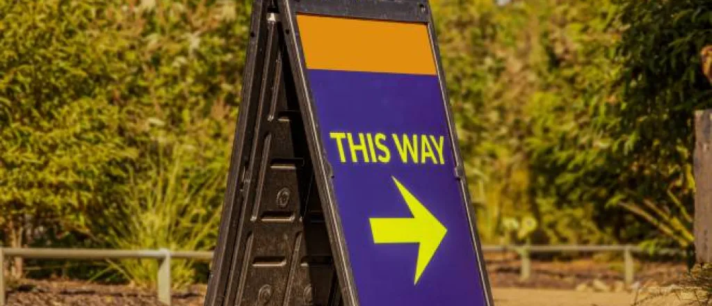 A large blue and orange A-frame sign with a printed message in yellow that says "THIS WAY" and an arrow pointing right.