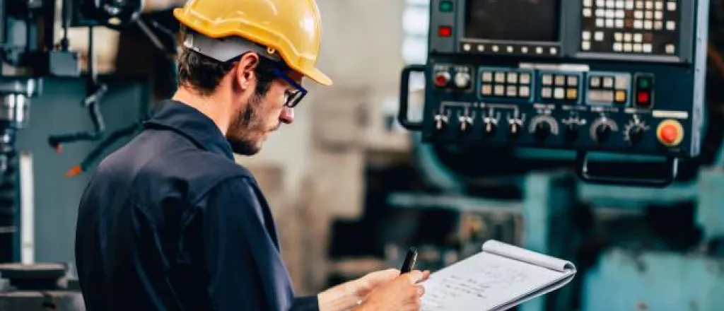 A team member wearing a blue jumpsuit and a yellow hard hat stands next to an automation machine, writing on a notepad. A team member wearing a blue jumpsuit and a yellow hard hat stands next to an automation machine, writing on a notepad.