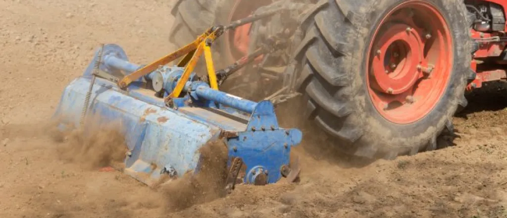 A large red tractor with a blue rotary tiller attachment The tiller is kicking up clouds of dirt in a field.