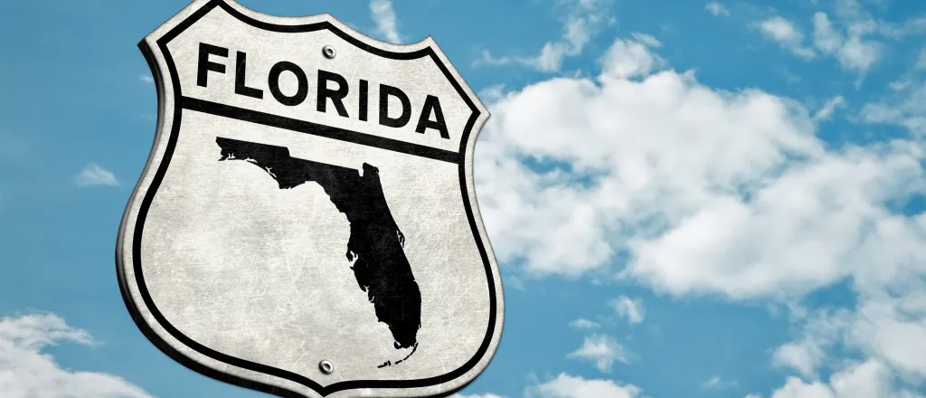 Road sign with the world "Florida" and a silhouette of the state against a clouded sky background.