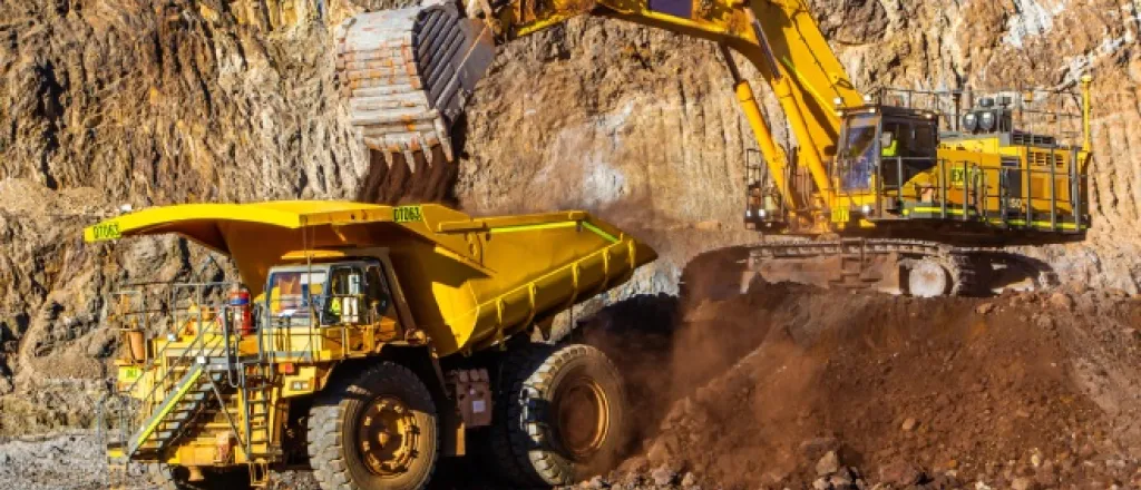 Two mining vehicles work together at a mining site. A digger on top of a small hill loads a dump truck with material.