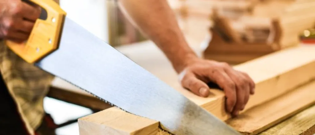 A close-up of a man using a large hand saw to cut a long piece of wood. He is holding the wood in place while he saws.