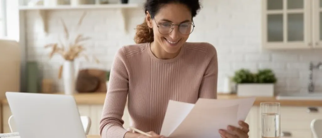 A female homeowner sitting at the dining table reading through printed tax documents. She has a laptop in front of her.