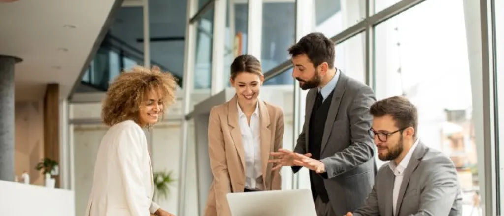 Four employees in business attire are clustered together around a laptop, looking at the screen.