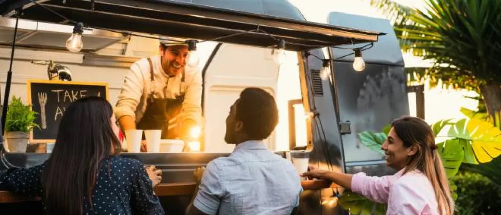Three adults stand at the window of a food truck, and a man inside the food truck smiles at them and wears an apron.